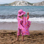 A child wearing a UV Protective Sun Hat Pink Starfish, stands on a sandy beach, back to camera, looking out at the waves of the sea.